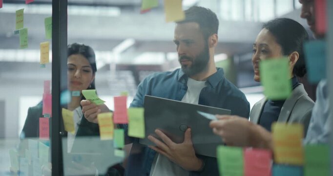 Employees Use Sticky Notes on a Glass Wall to Map Out Product Features and Steps in a Project. Colleagues Interact, Sharing Insights, Reordering notes, Using Laptop Computer and Building a Visual Plan