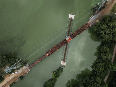 Aerial view of a rusty train trestle swing bridge cutting across a murky river, flanked by trees and power lines. Appleton, Wisconsin, United States.