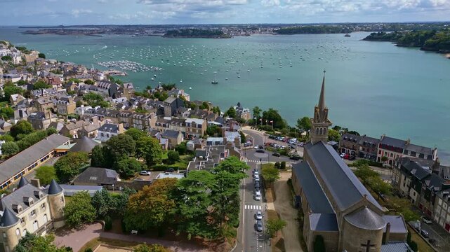 Dinard coastal town panorama with historic buildings and Church of Our Lady of the Emerald, boats on the Rance estuary and distant Saint Malo under a bright summer sky, drone pulling away