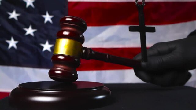 Brown wooden ceremonial gavel hammer hitten on wooden podium by a judge justice hand while a holy cross of faith moves in the foreground and American United States flag is in the background.