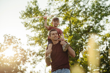 Cheerful father carries his joyful son on his shoulders in park at sunset. Boy is holding a toy airplane and smiling as play with dad outdoors. Child care and happy fatherhood concept. Space for text