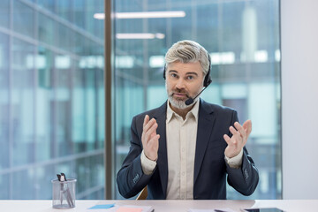 Mature man with a headset talking, gesturing with hands, and providing support or information during a call center shift or virtual consultation in a modern office