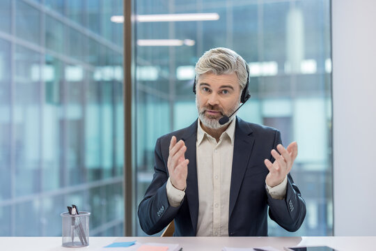 Mature man with a headset talking, gesturing with hands, and providing support or information during a call center shift or virtual consultation in a modern office