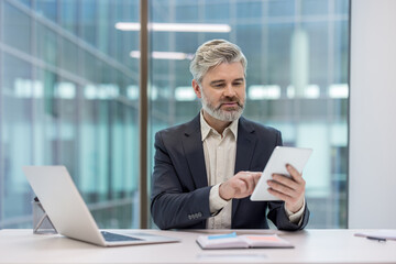 Mature businessman using a digital tablet at his office desk, focusing on the screen, reflecting modern technology integration in corporate environments