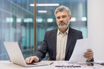 Confident mature businessman with gray hair and beard working at his office desk, reviewing documents and typing on a laptop, portraying professionalism and corporate success