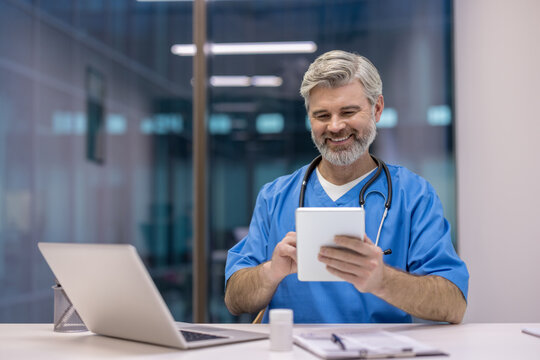 Experienced male physician in blue scrubs smiling at desk while using a digital tablet in a modern clinic, conveying confidence, telehealth readiness and friendly patient-focused care