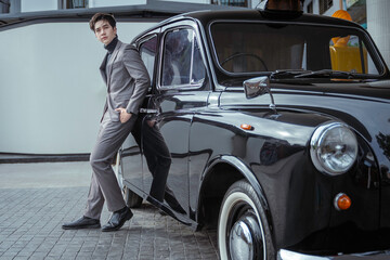 Handsome young man in a gray suit posing beside a vintage black car outdoors. An elegant fashion portrait with the retro car.