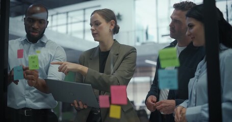 Diverse Creative Team Gathers Around a Glass Wall in Office, Using Sticky Notes to Sketch Out Ideas and Define Project Steps. Each Participant Shares Input while Adjusting a Visual Plan - Powered by Adobe