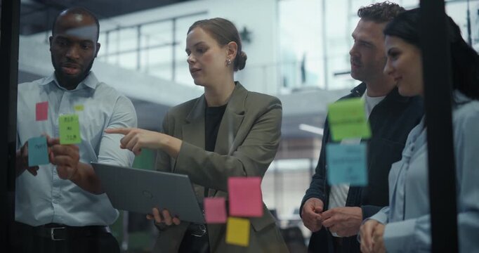 Diverse Creative Team Gathers Around a Glass Wall in Office, Using Sticky Notes to Sketch Out Ideas and Define Project Steps. Each Participant Shares Input while Adjusting a Visual Plan