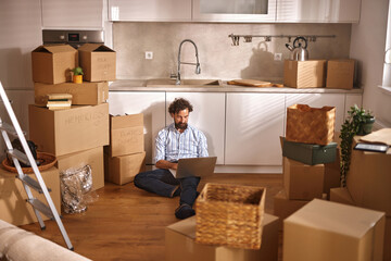 A man sits on the floor of a newly moved-in kitchen, surrounded by packed boxes and moving supplies. He focuses on his laptop, adjusting to the changes in his life.