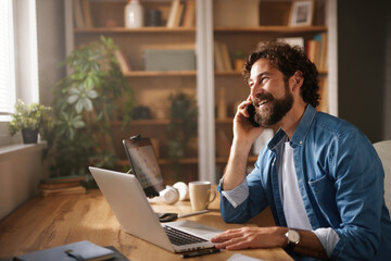 A man with a beard sits at a wooden desk in his home office, engaged in a phone conversation while typing on his laptop. Bright light streams through the window, illuminating the room.