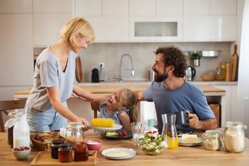 A warm breakfast gathering features a woman serving corn to her child while a man enjoys a drink, creating a joyful family moment in their bright kitchen.