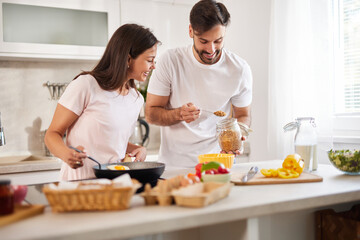 In a sunny kitchen, a couple happily cooks together, measuring grains from a jar while smiling. Fresh vegetables and fruits are scattered on the counter, creating a cheerful ambiance.