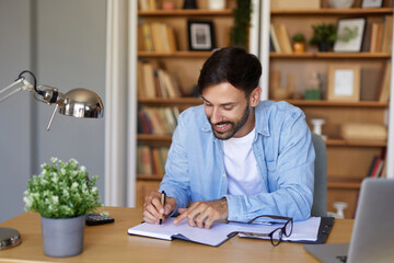A man sits at a desk in a cozy home office environment, happily writing in his notebook. Shelves filled with books and a small plant add to the tranquil atmosphere.