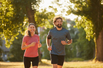 Two individuals run together in a park during sunset, surrounded by trees and vibrant greenery,...