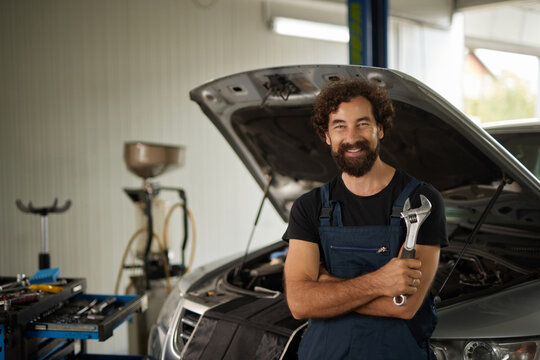 A skilled car mechanic stands inside a well-equipped repair shop, smiling while holding a wrench. The car hood is open, revealing the vehicle's engine, showcasing a busy work environment.