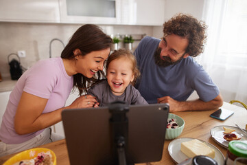 Obraz premium Family members gather around a tablet at the kitchen table, sharing a joyful moment while enjoying breakfast and video chatting with loved ones, creating warm memories together.