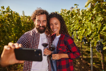 A couple stands in a lush vineyard, smiling and holding glasses of red wine while taking a selfie. The sun shines brightly overhead on a beautiful day, highlighting the grapevines.