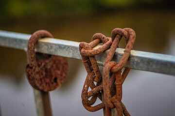 Close-up of an old, corroded chain and lock hanging on a weathered metal railing, with a blurred river in the background.
