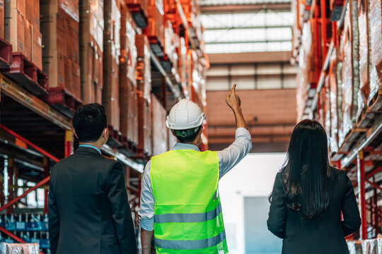 manager and a woman in suits, stand with a warehouse worker in a hard hat and safety vest. The worker points up toward the tall, stocked shelves as he talks to them.