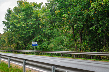 Fototapeta premium Empty highway bordered by metal guardrails and dense green forest, with a blue Murjāni city sign indicating entry into the town.