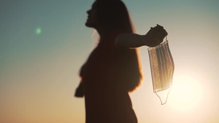 Woman holding mask at sunset with arms outstretched silhouette against sun and sky conveying freedom and relief after pandemic emphasizing health and outdoor recovery under warm sunlight - Powered by Adobe