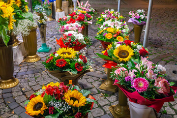 Obraz premium Flower bouquets with yellow sunflowers, roses and gerberas displayed in decorative stands on cobblestone street outside florist shop.