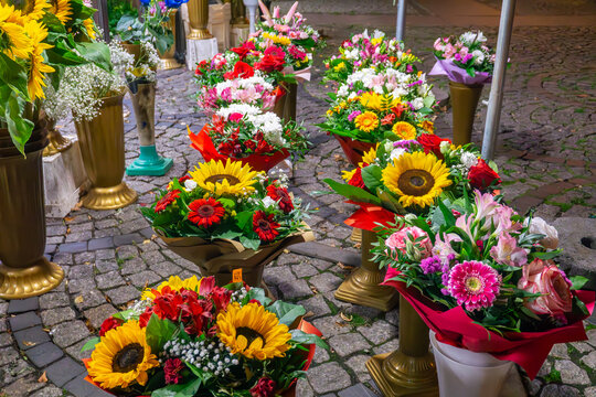 Flower bouquets with yellow sunflowers, roses and gerberas displayed in decorative stands on cobblestone street outside florist shop.