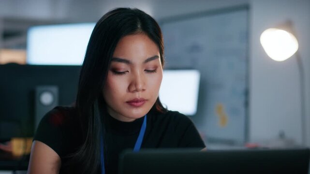 Professional Female Asian Software Engineer, Programmer working on laptop, looking at screen focused on writing code, Debugging Applications, developing app, Reviewing code working overtime in office.
