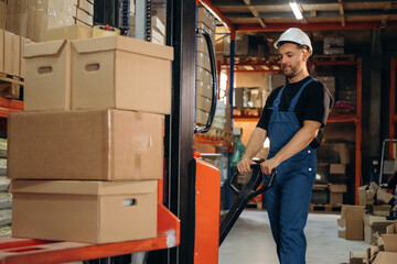Transporting the boxes by using forklift. Man in uniform is working in the storage with packages for shipment