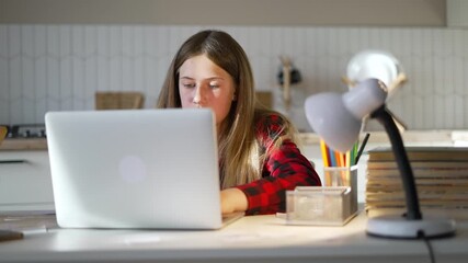 Studying on laptop at desk teenager girl completes homework under lamp with notebook while learning online study routine shows focused student engaged in education and school task concentration