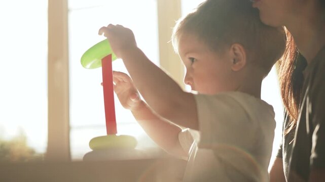 Mother plays with toddler stacking ring toy by sunlit window with parent guidance and child focus on learning and play fostering bonding development and fine motor skill growth in home environment