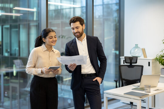 Fototapeta Two business colleagues discussing financial documents and using a digital tablet, standing together in a contemporary office environment, symbolizing teamwork and professional partnership