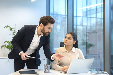 Professional manager guiding a focused female employee in a modern office, reviewing a business project on a laptop while collaborating and problem-solving