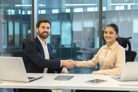 Diverse business professionals shaking hands at an office desk, symbolizing a successful deal, partnership, and agreement, reflecting collaboration and teamwork in a corporate environment - Powered by Adobe