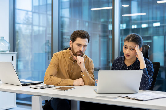 Two diverse business colleagues are concentrating and discussing a problem at their desk in a modern office, working together to find a solution on a laptop