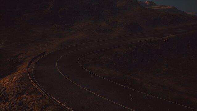 A serene winding road cuts through rugged mountain landscape as dusk approaches. The fading light casts shadows on the terrain, creating a tranquil yet mysterious atmosphere.