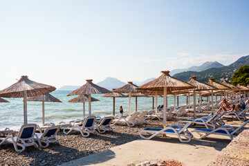 Sunny Mediterranean beach with rows of straw parasols and empty sun loungers by the sea. Perfect holiday and summer vacation atmosphere.