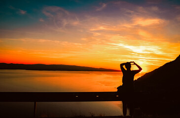 Fototapeta premium Silhouette of a person making a heart shape with hands at sunset, overlooking calm sea and mountains, warm sky colors creating romantic and peaceful mood