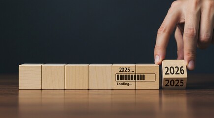 In the image, a person's hands are shown adjusting wooden blocks, with the numbers on the blocks indicating the year is changing from 2025 to 2026. A loading bar is visible on one of the blocks, servi