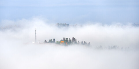 Picturesque view of hillside adorned with vibrant autumn foliage, ranging from rich greens to fiery reds and golden yellows. Wisps of fog drift across landscape under bright blue sky.
