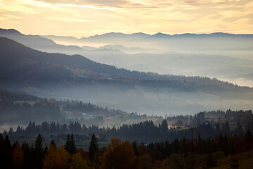 Misty autumn morning in mountain valley. Layers of hills bathed in soft light, with patches of sunlight breaking through fog, illuminating autumn foliage. Interplay of light and shadow.