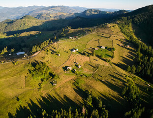 Aerial view of rural landscape at sunset, with rolling hills and scattered farmhouses. Winding paths and patches of dense forest add texture and depth to the scene.