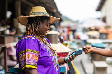 Contactless transactions, small business owner, SME, local traditional African market woman accepts payment via a mobile point of sale, POS system at an outdoor market stall.