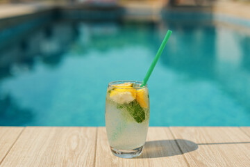 A cold drink with a green straw and lemon slices is placed on a wooden deck next to a swimming pool. The image evokes feelings of summer, holidays, and relaxation.