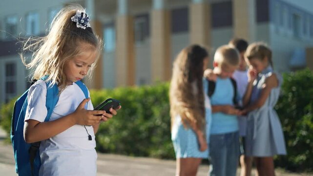 Girl uses smartphone alone near school while group of child peers gather nearby with backpack and phone causing social separation and subtle bullying and loneliness in sunny outdoor playground setting