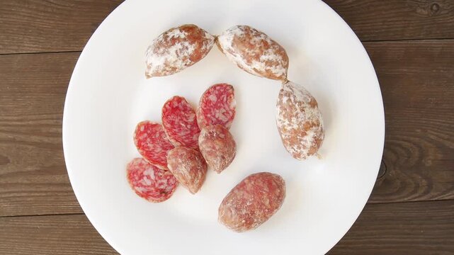 Small salami sausages, both whole and sliced, rotating on a white plate over a wooden table.