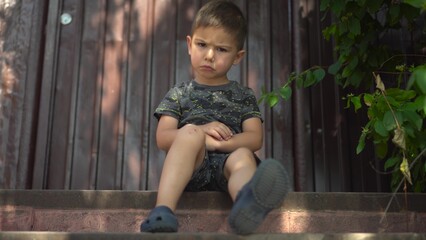 Child is angry about a bruised knee while sitting on the porch in dark clothes on a hot summer day