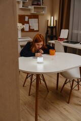 A teenage girl studies in the living room during evening hours, drinking coffee while focusing on homework. Captures authentic student life and modern study routines