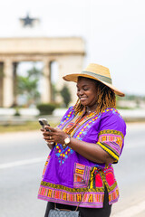 Fototapeta premium Smiling African Ghanaian woman in traditional purple dashiki and straw hat uses smartphone outdoors for video calls, online shopping, digital banking, social media and internet browsing.